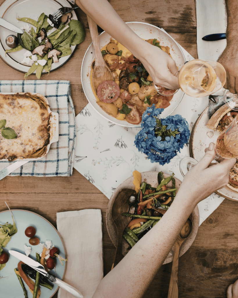 An overhead view of a dining table set with various dishes, including a salad, a vegetable medley, and a lasagna. Hands are reaching for food and glasses of drink, with a blue flower centerpiece adding color.