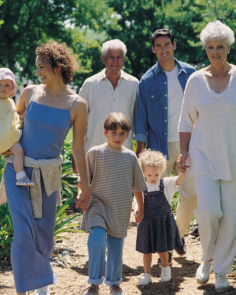 A diverse family walking together in a park, smiling and enjoying each other's company. The group includes a grandmother, a father, a mother holding a baby, and three children.