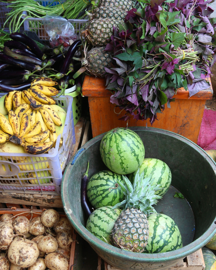 A variety of fresh fruits and vegetables at a market, including bananas, watermelons, pineapples, eggplants, and leafy greens, displayed in baskets and containers.