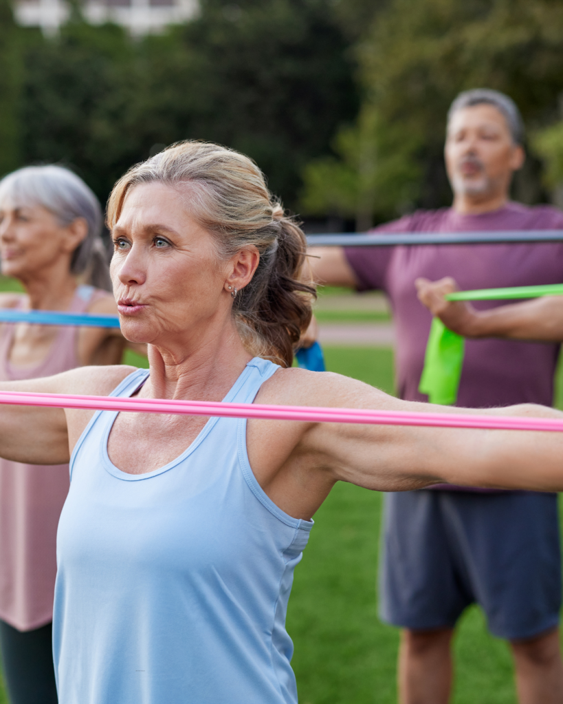 A group of older adults performing resistance band exercises in a park, with a focus on a woman in a blue tank top engaging her upper body.