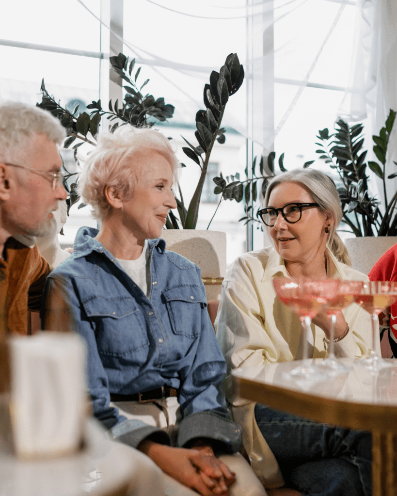 A group of four older adults sitting together in a stylish cafe, engaged in conversation, with drinks on the table and tropical plants in the background.