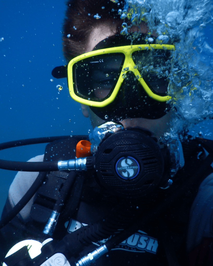 A diver wearing yellow snorkeling goggles and a black diving mask, submerged underwater with bubbles around.