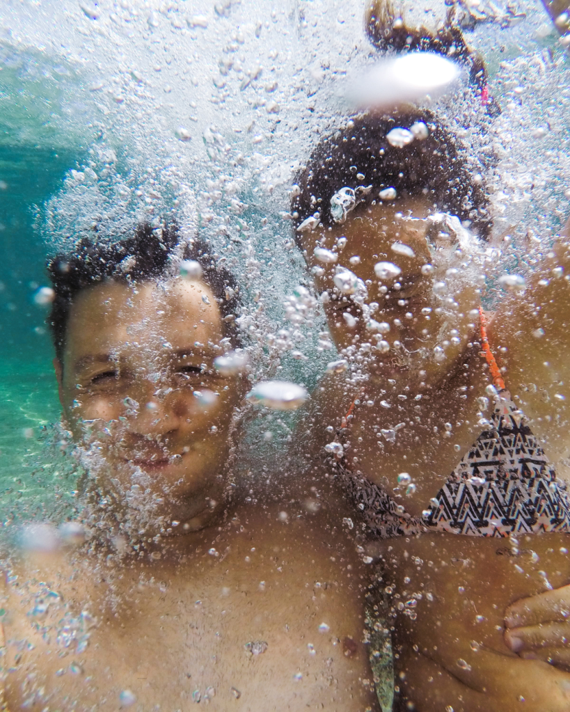 Two people taking a selfie underwater, surrounded by bubbles and clear blue water.