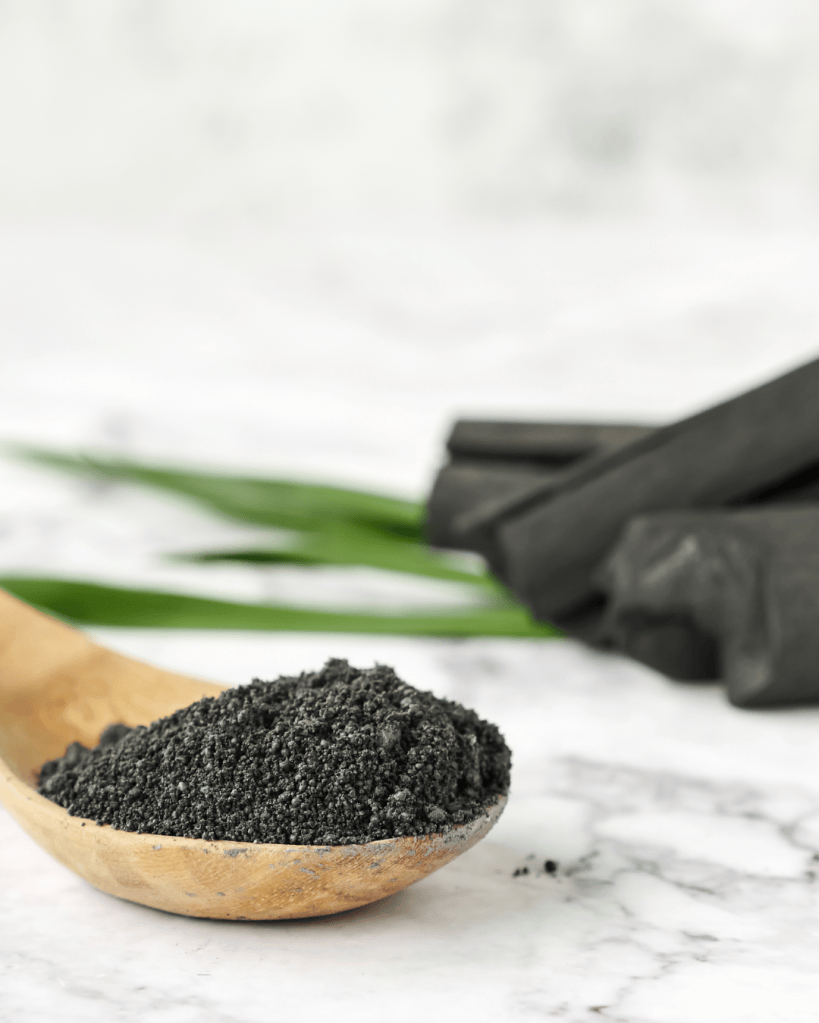 A wooden spoon filled with black powder, with pieces of charcoal and green leaves in the background.