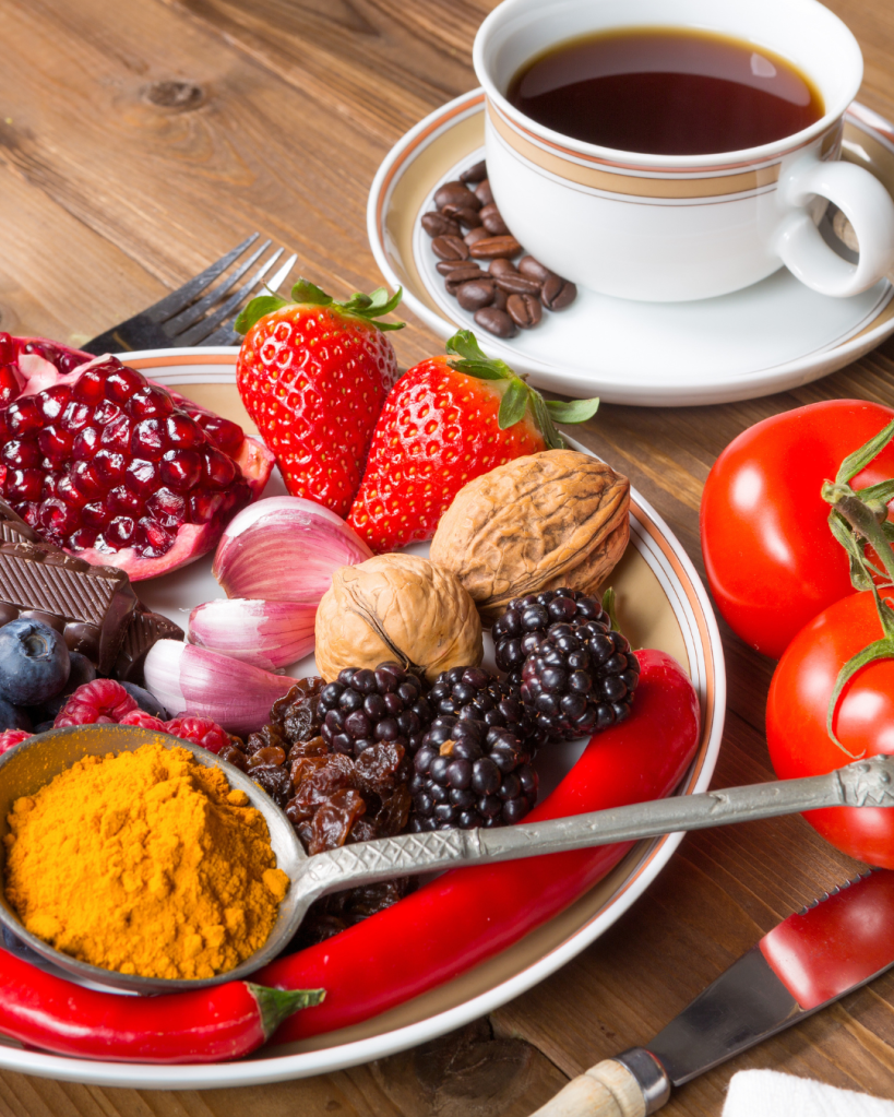 A colorful assortment of fresh fruits and spices on a plate, including strawberries, pomegranate, walnuts, and blackberries, with a side of chili peppers. A cup of coffee and some chocolate are also visible in the background.