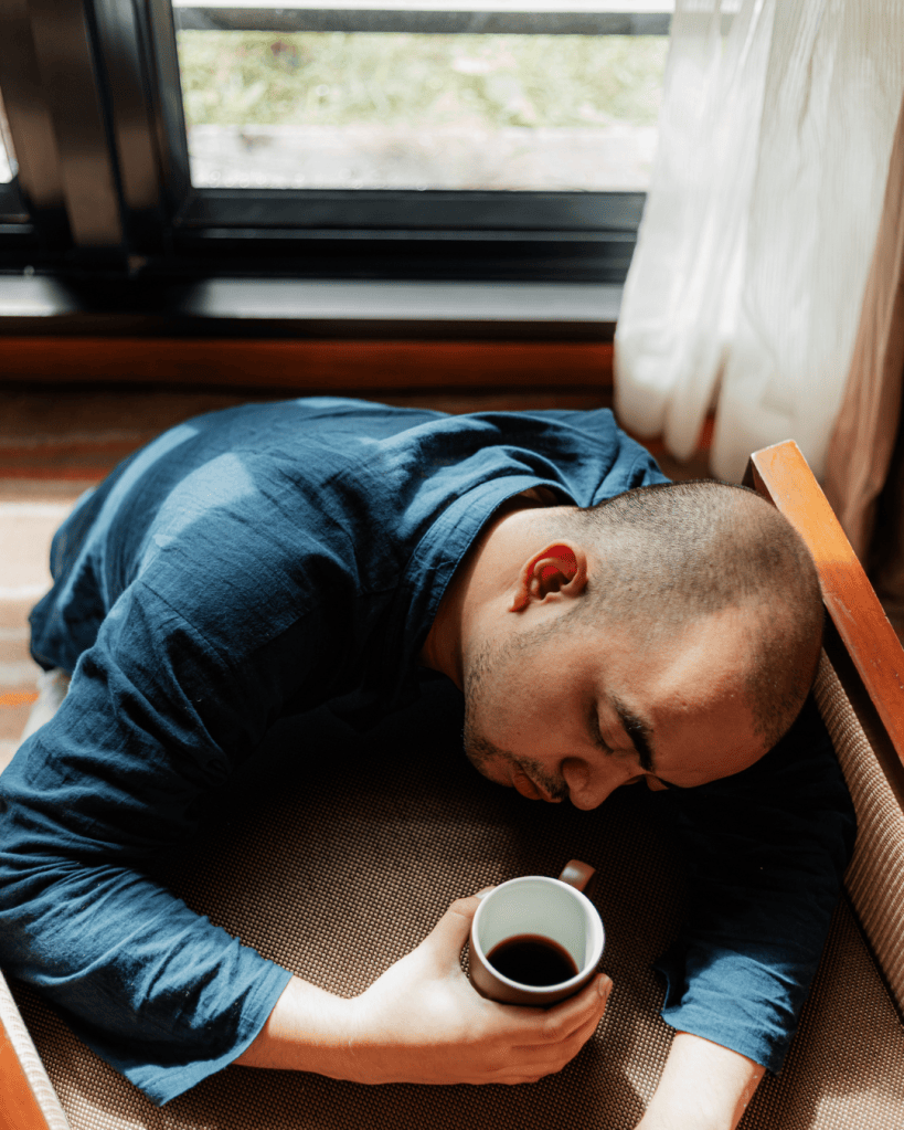 A person resting with their head down on a chair, holding a cup of coffee, with sunlight streaming through a window.