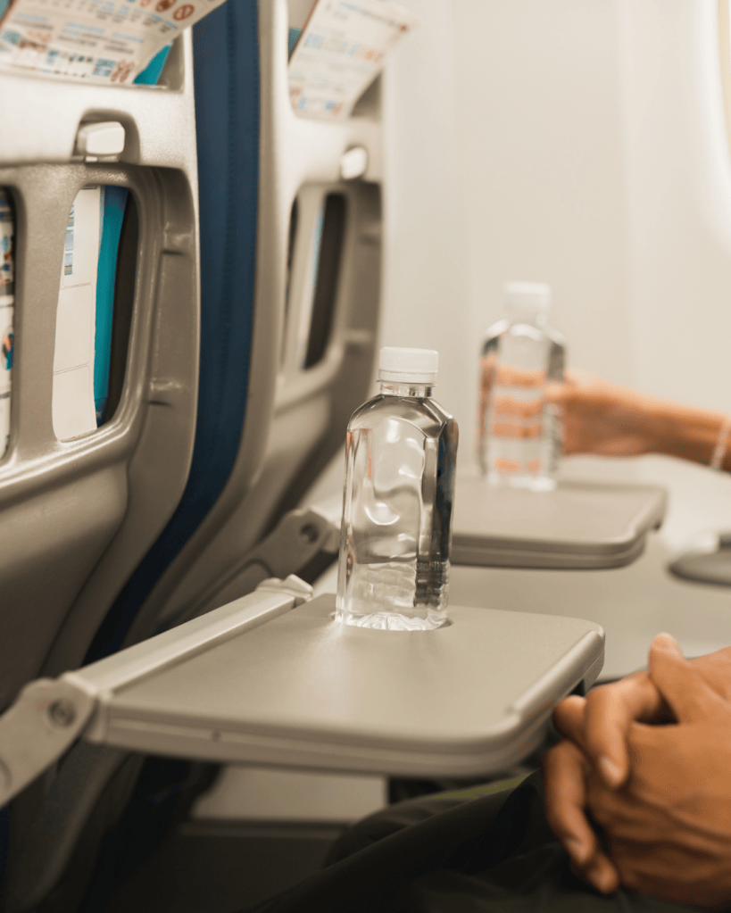 Two airplane seats with a water bottle on a tray table, one passenger's hands are resting on their lap.