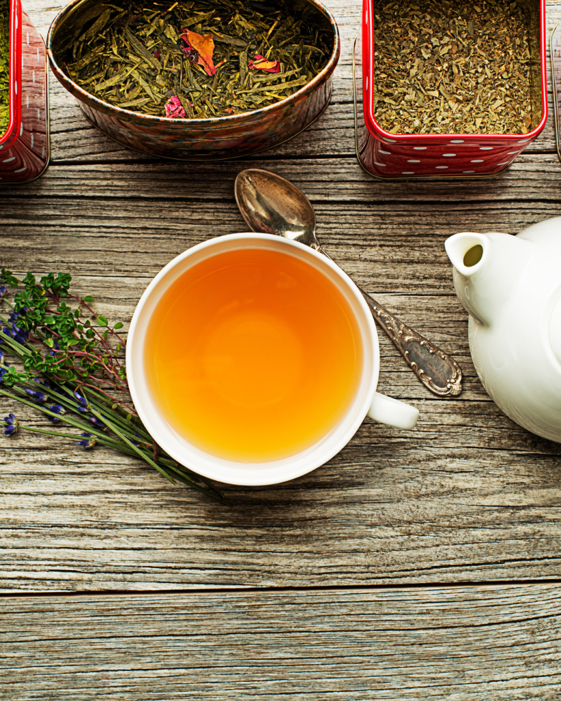 A cup of brewed tea on a wooden table, accompanied by a vintage spoon and a white teapot, with various tea leaves in decorative tins nearby.
