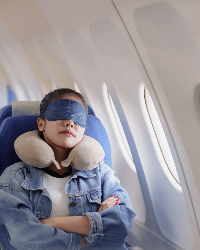 A woman sleeping on an airplane, wearing a sleep mask and a neck pillow, with her arms crossed and a relaxed expression.
